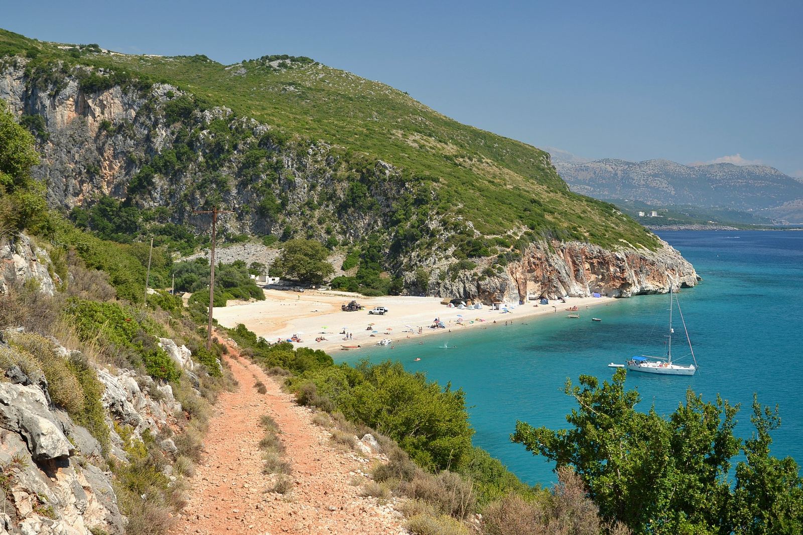 Footpath along the rocky coast to the white beach of Gjipe with turquoise water and a sailing boat