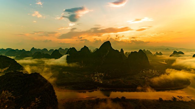 Sunrise over the Li River: dark karst peaks rise from the morning mist under a golden sky, with a village and the river in the valley