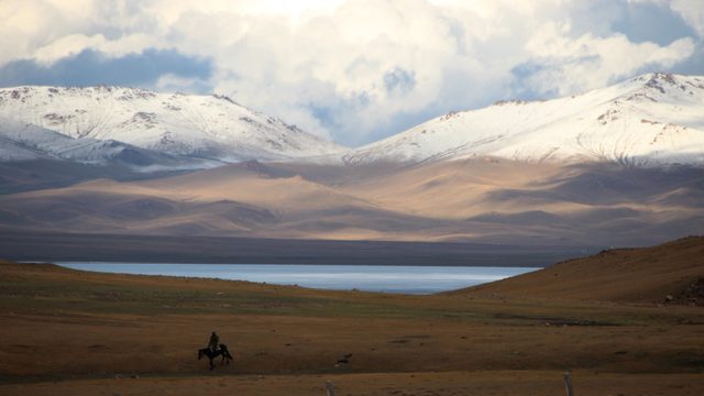 Rider on horseback on the steppe by Song-Kul lake, with snow-capped peaks under a cloudy sky