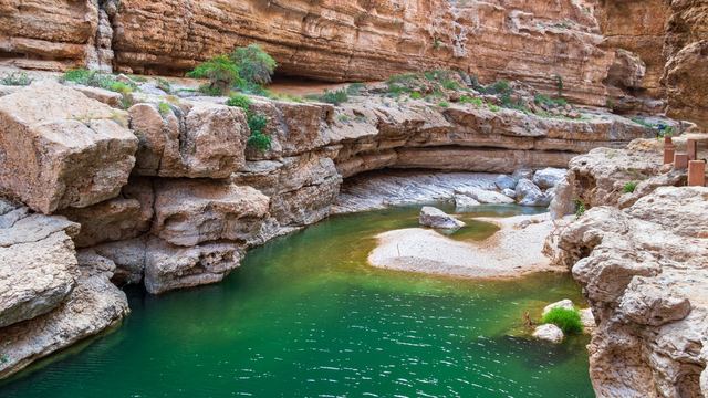 Emerald-green pool between layered limestone walls and boulders in the Wadi Shab gorge
